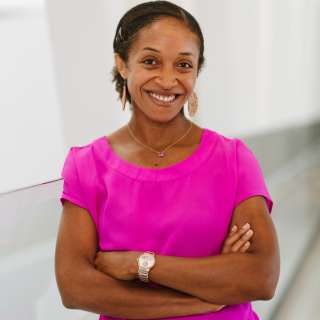Aisha Lofters, seen from the waist up, wearing a pink blouse with arms crossed and smiling