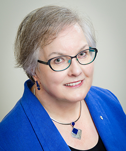 Anne Martin-Matthews, seen from the shoulders up, wearing a blue blazer, necklace, glasses, and short grey hair