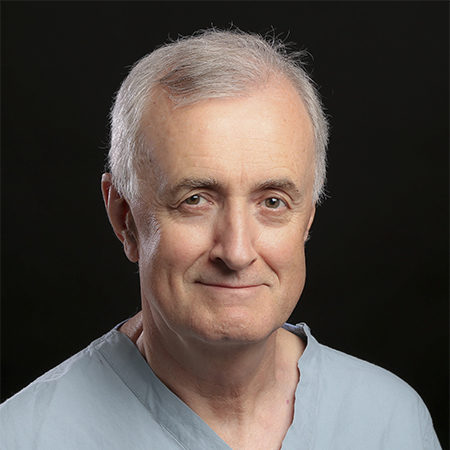 John Semple, seen from the shoulders up, wearing green scrubs, short grey hair, and smiling