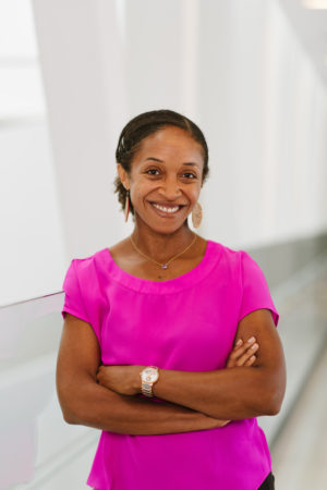 Aisha Lofters seen from the waist up, wearing a pink blouse with arms crossed and smiling