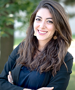 Razan Rawdat, seen from the chest up, wearing a black blazer, ornate earrings, long brown hair, and smiling