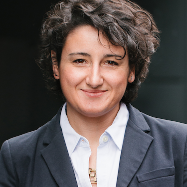 Arlinda Ruco, seen from the shoulders up, wearing a blue suit, short black curly hair, and smiling