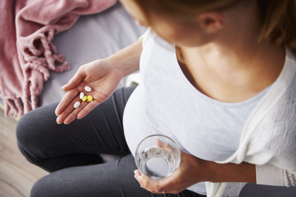A pregnant woman seen from above, holding a handful of various pills in one hand and a glass of water in the other