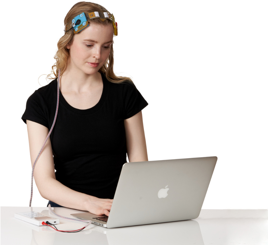 a woman wearing electronic sensors on her head sits and works on a laptop connected to the sensors